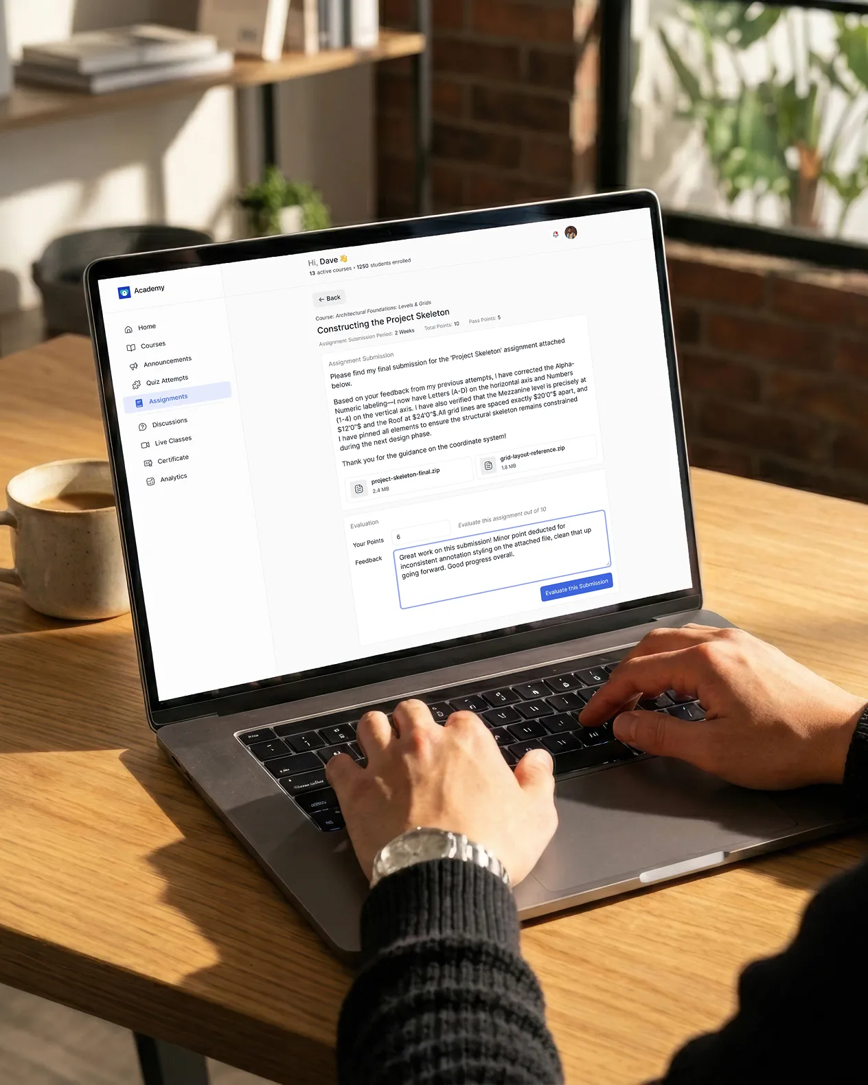 A person on laptop, writing assignment feedback after evaluating a submission on a brown table and a coffee mug behind the laptop in a room with a window and a bookshelf slightly visible and