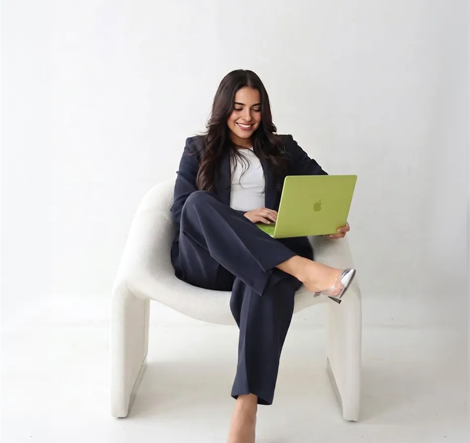 A woman in a navy blazer sits cross-legged on a white sculptural chair, smiling while working on a green MacBook against a clean white background.