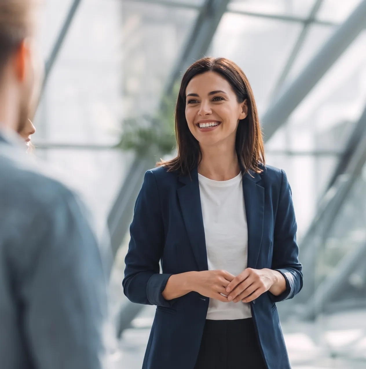 Smiling businesswoman in a navy blazer talking with a male colleague in a bright, glass-walled office.
