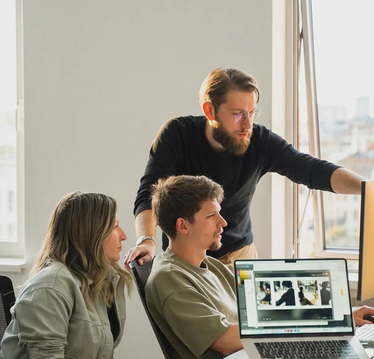 A team collaborates in a modern open-plan office, with a colleague pointing at a monitor while reviewing work together. A whiteboard behind them shows flowcharts and sticky notes organized under sections like 
